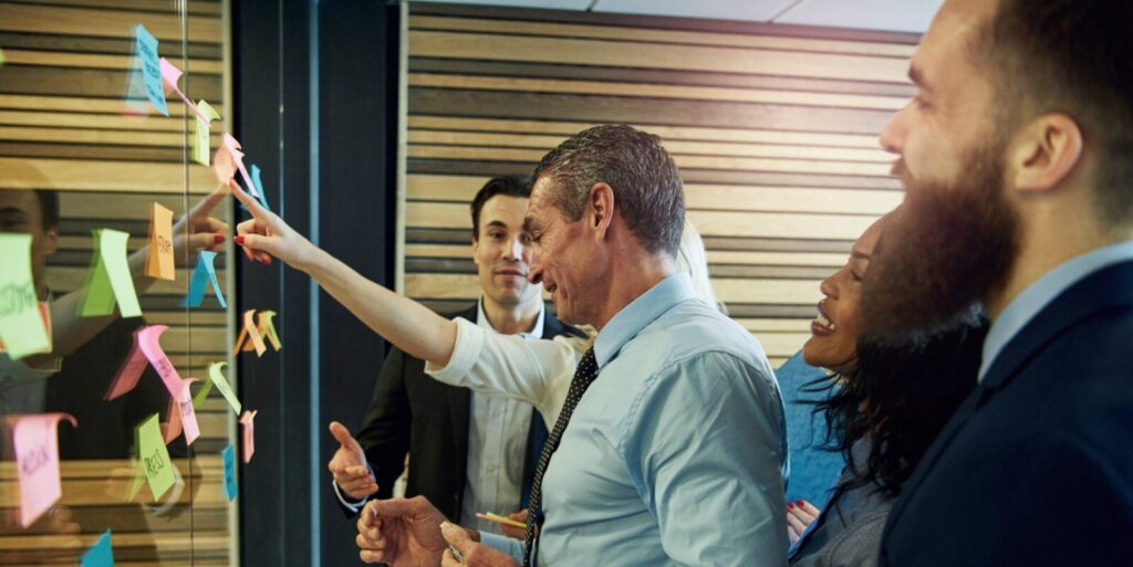 Four people in business attire stand by a glass wall with colorful sticky notes, discussing and pointing at the notes in a collaborative meeting.