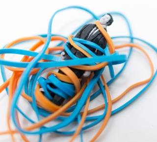 A small black object tangled in multiple orange and blue rubber bands on a white background.