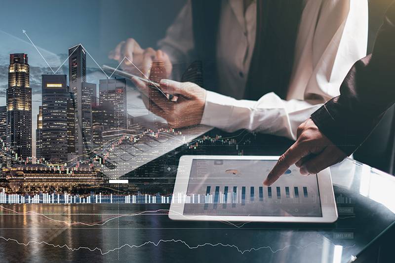 Two people work with a tablet displaying bar graphs, with a city skyline and stock market charts digitally overlaid on the image.