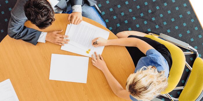 Two people sit at a round wooden table reviewing and signing documents, viewed from above. One person holds a pen while the other points at the paper.