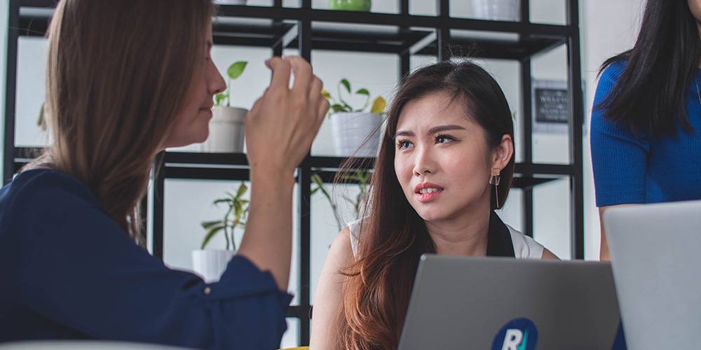 Two women are having a serious conversation in an office setting, with laptops and potted plants visible in the background.