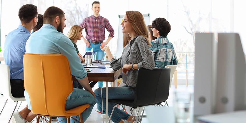 A group of people sit around a table in an office meeting while a man stands at the front of the room, speaking to them.