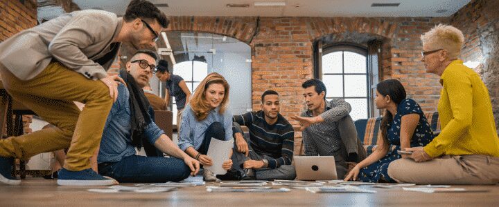 A group of people sit on the floor in a modern office, discussing papers and working on a laptop together.