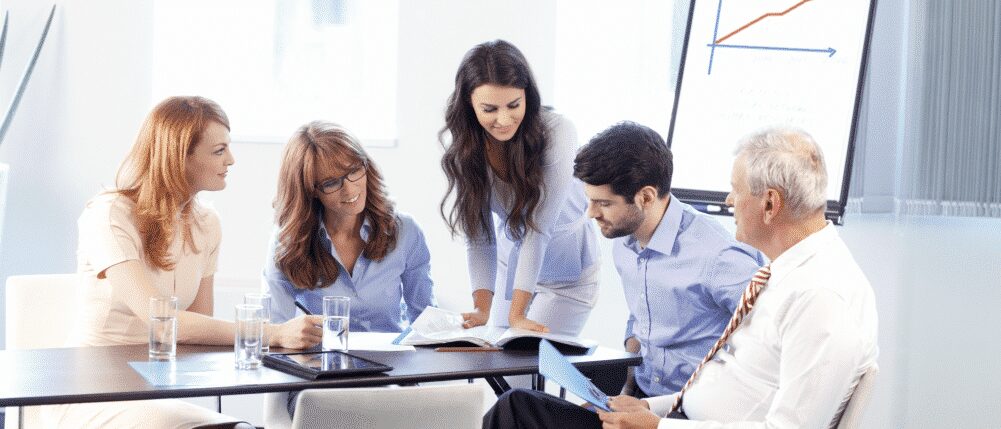 Five people in business attire sit and stand around a conference table, looking at documents, with a chart displayed on an easel in the background.
