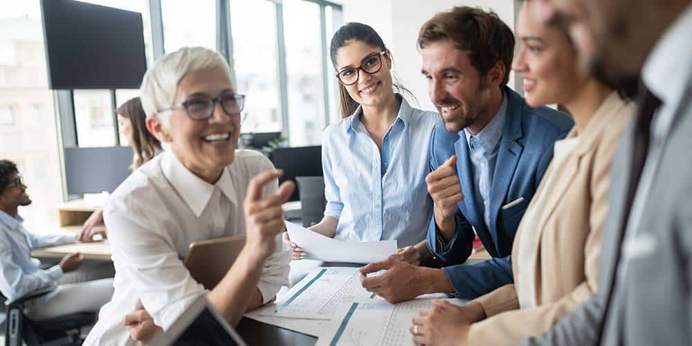 A group of professionals are gathered around a desk in a modern office, smiling and discussing documents during a meeting.