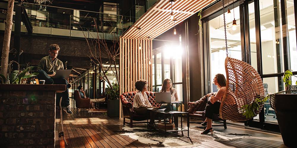 Four people sit and work in a modern, sunlit office with large windows, wooden floors, and a swing chair; one person sits on a ledge with a laptop.