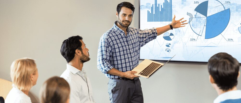 A man gives a presentation to a seated group, pointing at charts and graphs displayed on a large screen while holding a laptop.