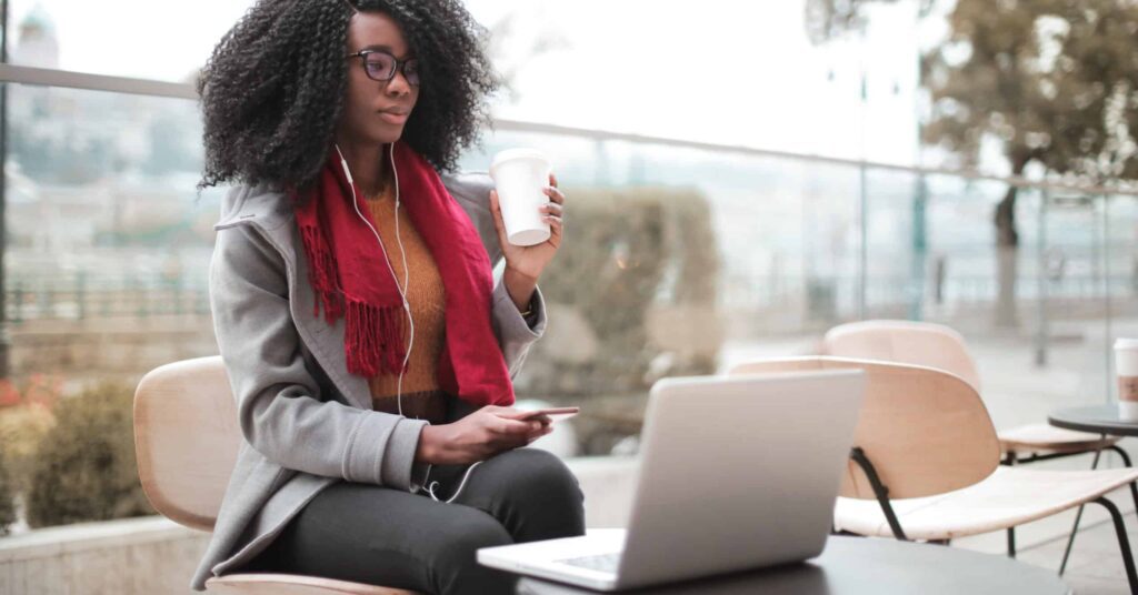 A woman sits outdoors at a table using a laptop, holding a coffee cup and listening to earphones, with empty chairs and trees in the background.