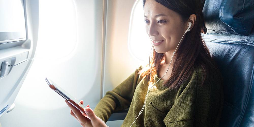 A woman sitting by an airplane window uses a tablet and listens to earphones, smiling while seated in an economy class seat.