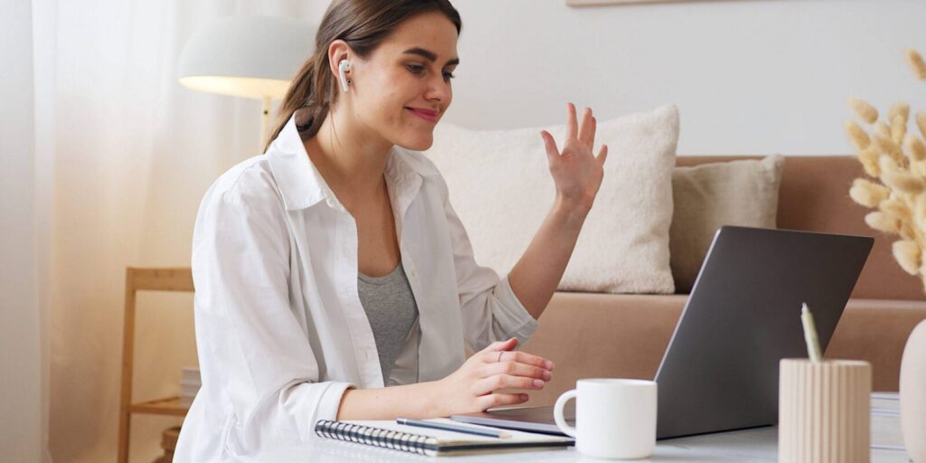 Woman wearing earbuds sits at a table, waving at a laptop during a video call. A notebook, pen, and coffee mug are on the table in front of her.