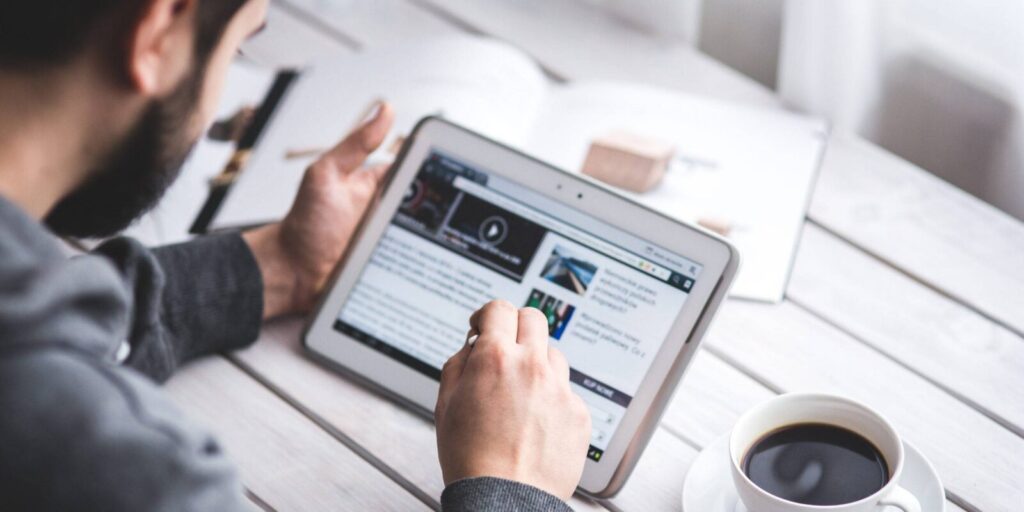 A person using a stylus to browse a tablet at a white desk, with an open magazine and a cup of coffee nearby.