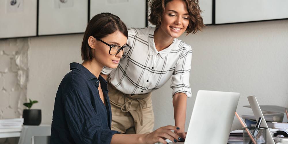 Two women working together at a desk, one sitting and typing on a laptop while the other stands beside her, both focused on the screen in a modern office setting.