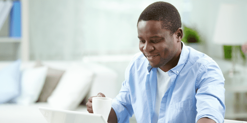 A man sits at a table using a laptop and holding a white mug, smiling, in a bright indoor setting.