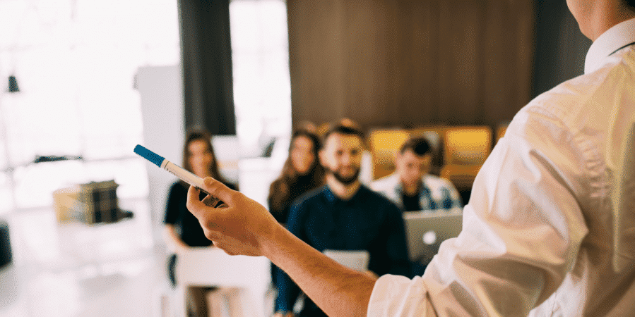 A person standing and holding a marker addresses a group of people seated and paying attention, with one using a laptop.