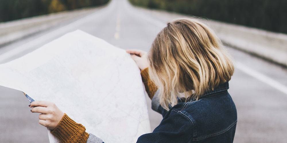 A person with blonde hair stands on an empty road while looking at a large unfolded map.