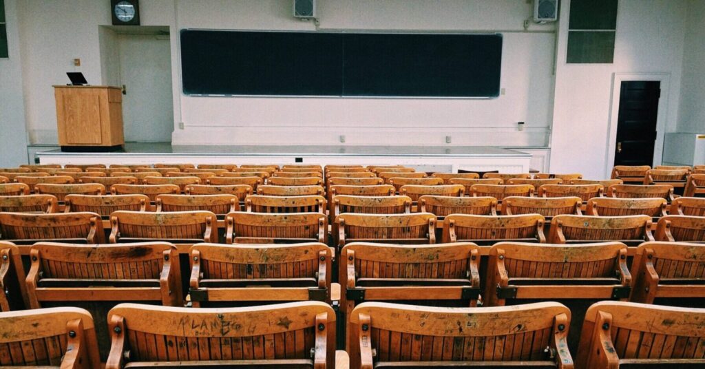 Empty lecture hall with rows of wooden seats facing a chalkboard, podium, and clock at the front of the room.