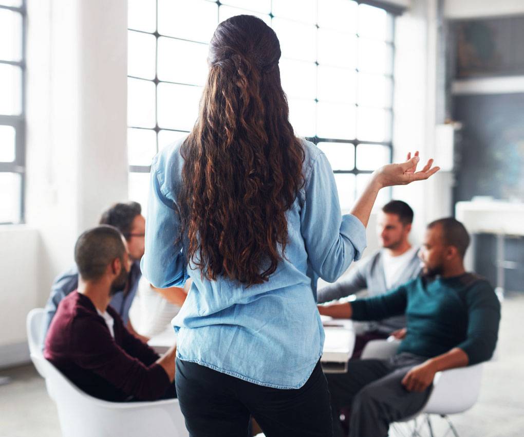 A woman stands addressing a seated group of four people in a modern, well-lit meeting room.