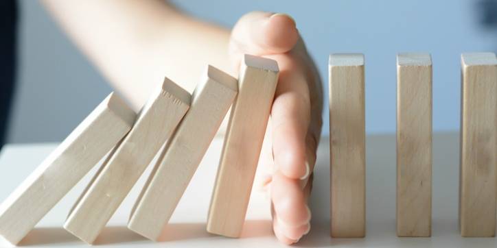 A hand stops a row of wooden dominoes from falling, interrupting the chain reaction.