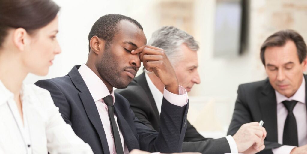 Four business people sit at a table; one man in a suit closes his eyes and pinches his nose, appearing stressed or tired, while others are focused on their work.