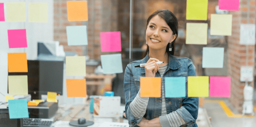 A woman stands in front of a glass wall covered with colorful sticky notes, holding a marker and smiling thoughtfully.
