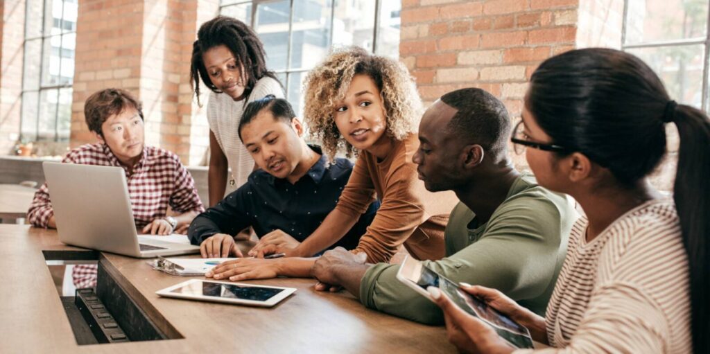 Six people sit around a table with a laptop and tablets, discussing documents in a modern office with brick walls and large windows.