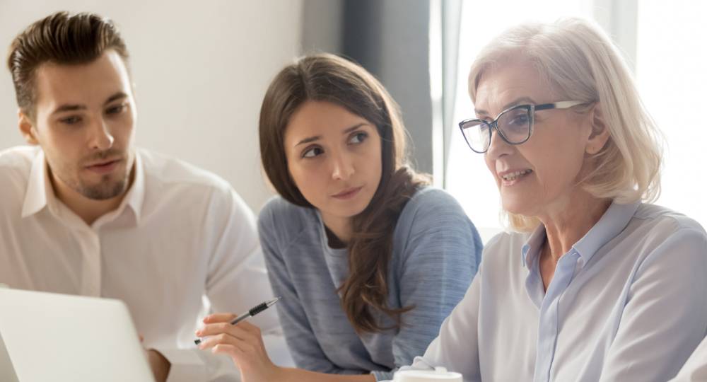 Three people sit at a table with a laptop, having a discussion. One woman is speaking while the other two listen attentively and take notes.