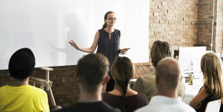 A woman stands in front of a whiteboard giving a presentation to a seated audience in a modern office setting with exposed brick walls.