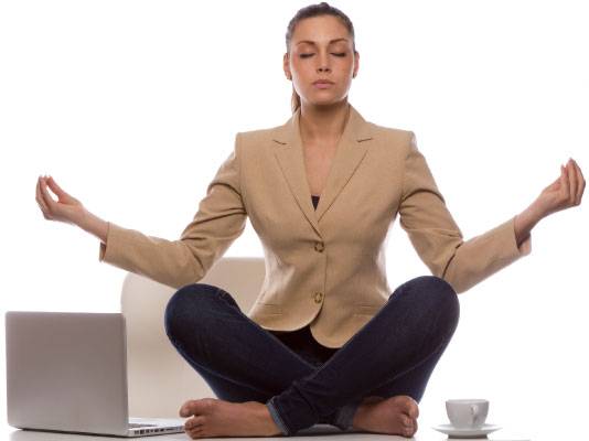 A woman in business attire sits cross-legged on a chair, eyes closed, meditating next to a laptop and a cup.