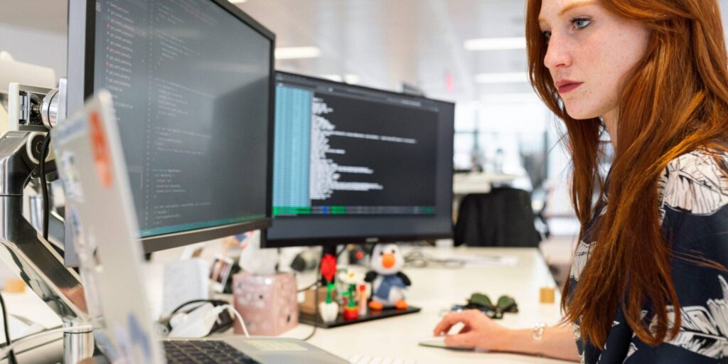 A woman works at a desk with two large computer monitors displaying code in a modern office setting.