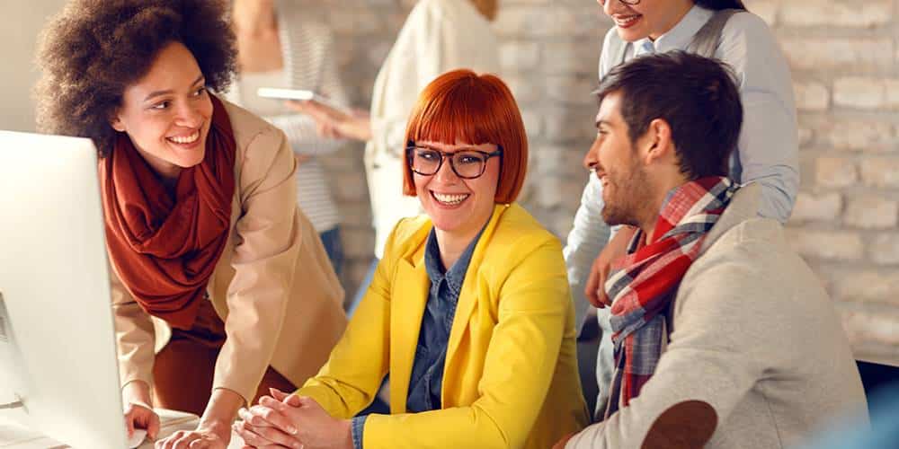 Four people are gathered around a computer in an office, talking and smiling. One person is seated wearing a yellow jacket, while the others are standing and engaging in conversation.