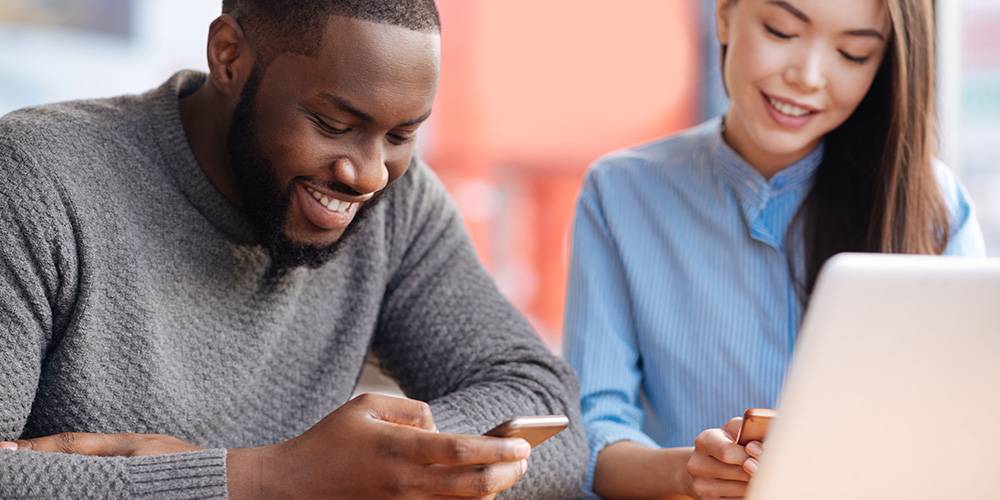 Two people sit at a table, both smiling while looking at their smartphones. One person also has an open laptop in front of them.