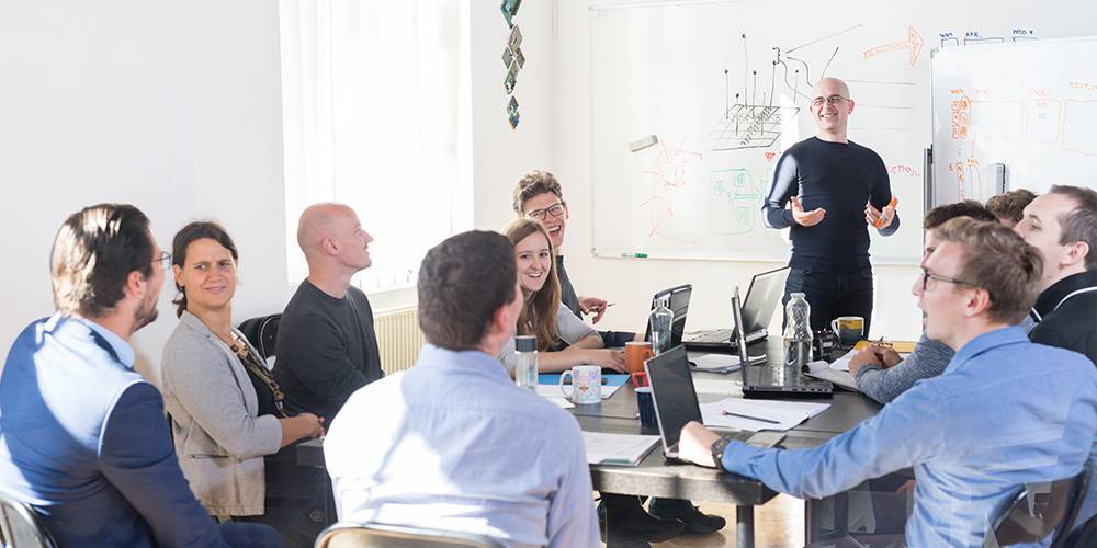 A group of people sit around a table with laptops and papers, listening to a man standing by a whiteboard giving a presentation in a bright office room.