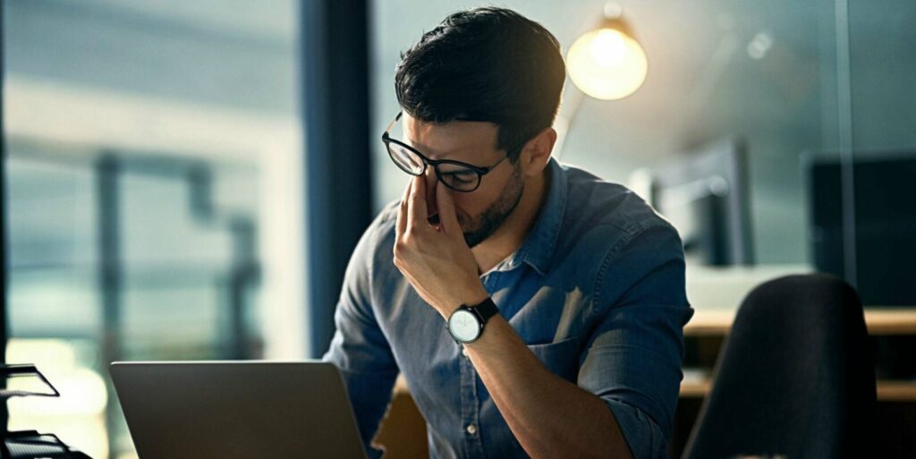 A person wearing glasses sits at a desk with a laptop, pinching the bridge of their nose, appearing stressed or fatigued in an office setting.
