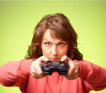 A woman in a pink hoodie holds a video game controller and looks forward against a green background.