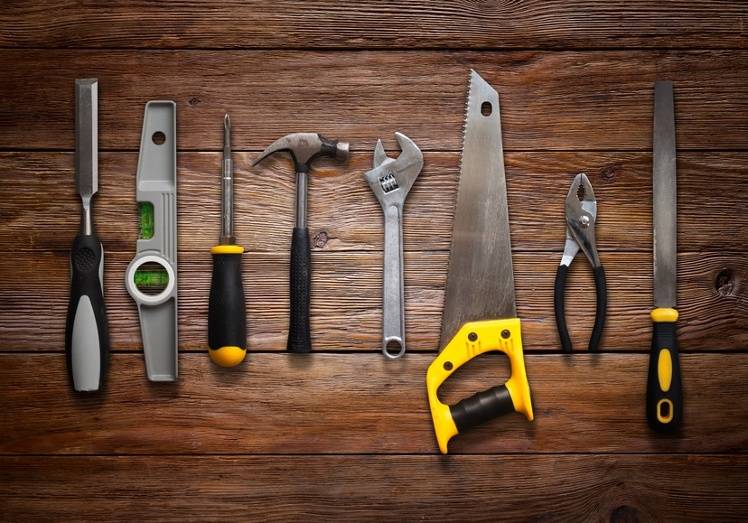 A row of eight hand tools, including a chisel, level, screwdriver, hammer, adjustable wrench, saw, pliers, and file, arranged on a wooden surface.