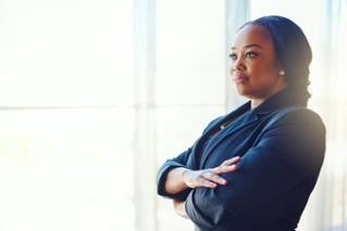 A woman in a dark blazer stands with arms crossed, looking out a bright window with a serious expression.