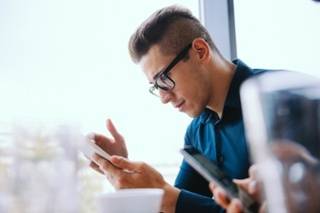 Man wearing glasses and a dark shirt looking at his smartphone, seated indoors at a table with blurred cups and another phone in the foreground.