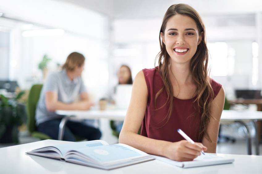 Young woman smiling and writing in a notebook at a desk with an open book; two people are talking in the background in an office setting.