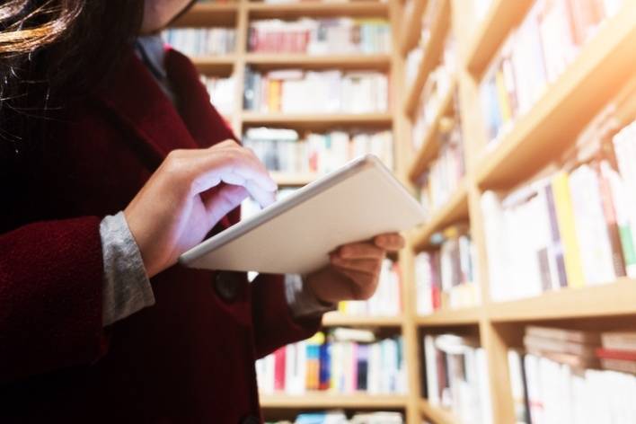 A person in a library browses a tablet, surrounded by bookshelves filled with books.