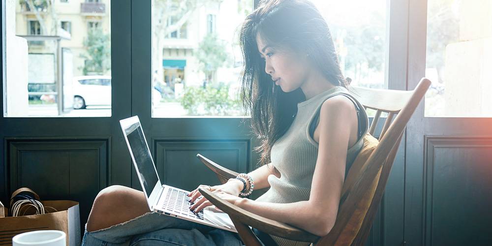 A woman sits in a wooden chair by a window, working on a laptop with sunlight streaming in behind her.