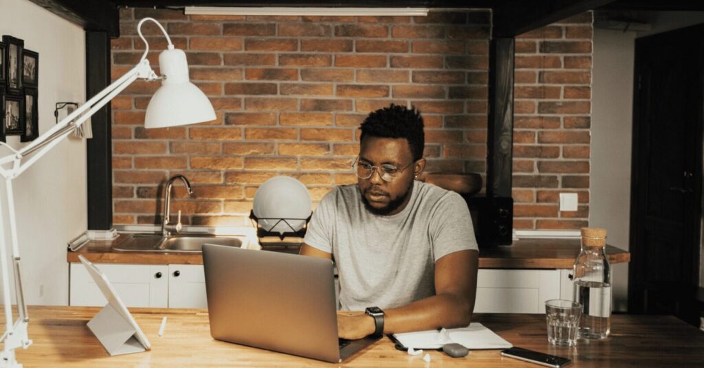 A person sits at a kitchen counter working on a laptop, with a tablet, notebook, glass of water, and water bottle nearby.