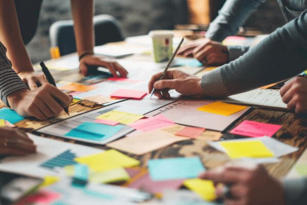 Several people are working together at a table covered with colorful sticky notes, papers, and pens, suggesting a collaborative brainstorming or planning session.