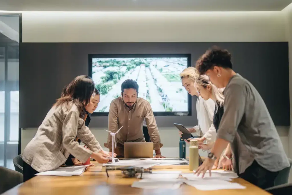 Six people stand around a table covered with papers and a model wind turbine, discussing work in a modern conference room with a screen displaying an aerial image in the background.