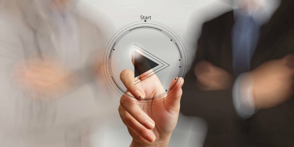 A hand presses a digital play button with the word “Start” at the top, while two blurred people in business attire stand in the background.