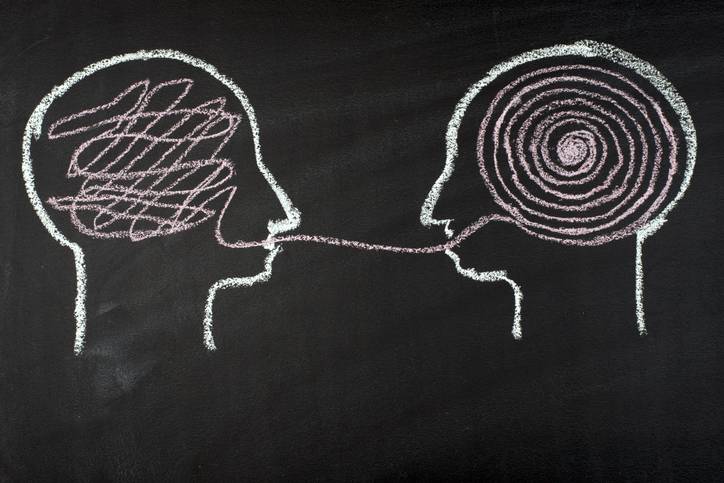 Chalk drawing of two heads facing each other; one with a scribbled brain and one with a spiral, connected by a line from one mouth to another, symbolizing communication or idea transformation.