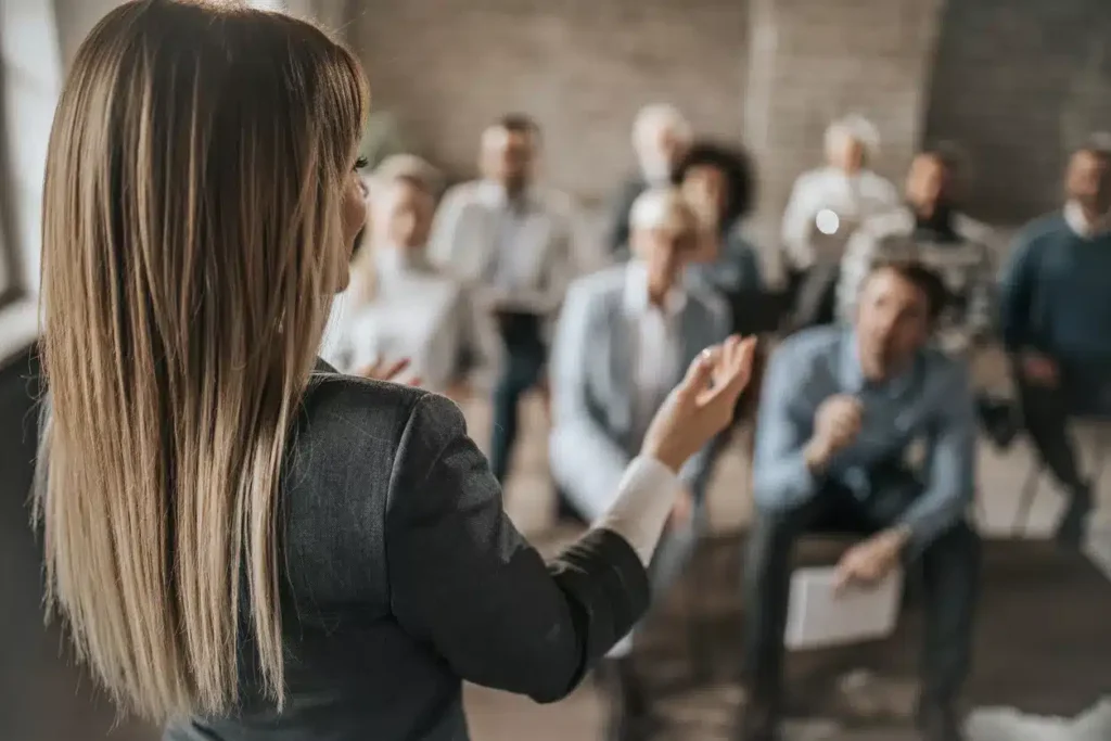 A woman speaks in front of a seated audience in a professional setting, with several people listening attentively.