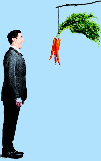 A man in a suit stands facing a bunch of carrots hanging from a stick suspended in front of him against a blue background.