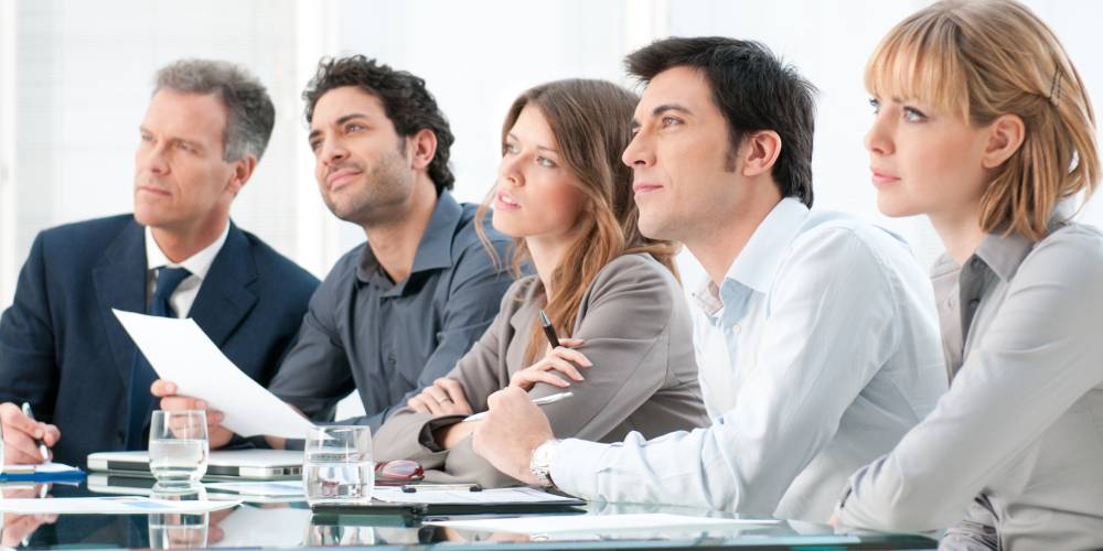 Five people in business attire sit at a conference table, attentively listening and looking in the same direction, with papers, glasses of water, and notebooks in front of them.