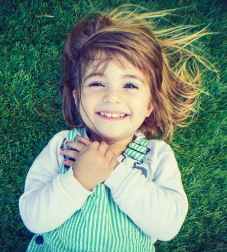 A young child with light brown hair lies on green grass, smiling up at the camera with hands crossed over their chest, wearing a striped overall and a white shirt.
