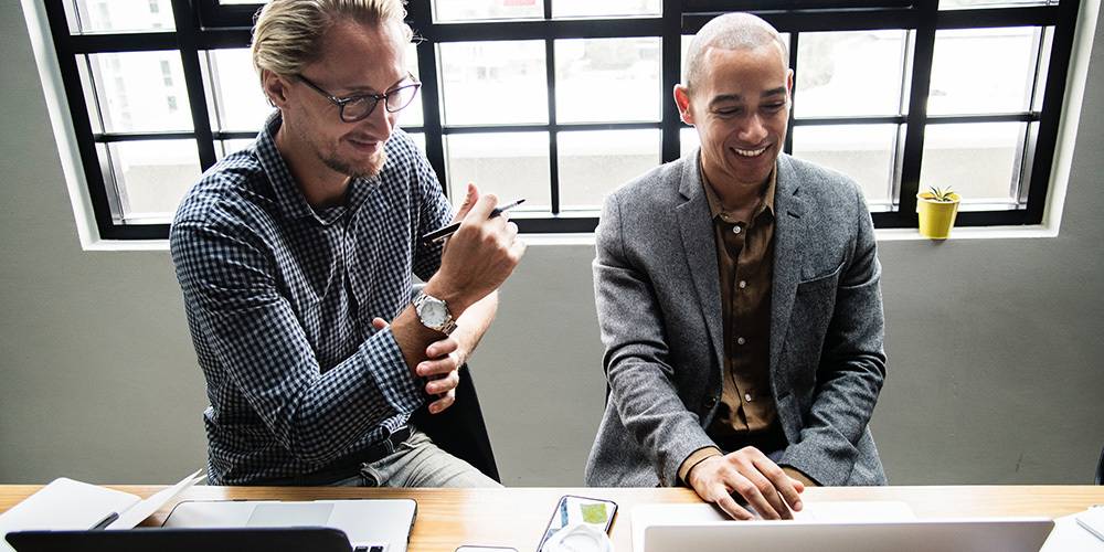 Two men sit at a desk with laptops, smiling and working together in a bright office with large windows.